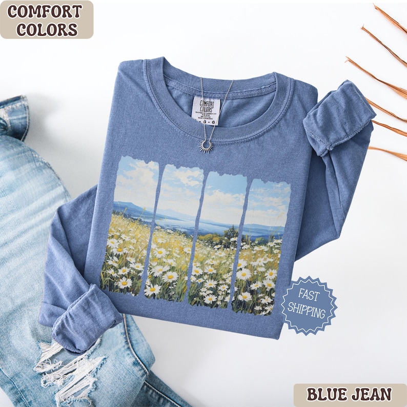 May include: A blue long-sleeve shirt with a landscape design featuring a field of white flowers and a blue sky. The shirt has the text "Comfort Colors" on the upper left and a silver necklace. The shirt is folded next to a pair of blue jeans.