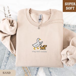 May include: Sand-colored sweatshirt with a crew neck, featuring an embroidered design of a duck walking a dog on a leash. The text "wag and waddle" is below the graphic. The words "Super Soft" are in a brown box in the upper right corner.
