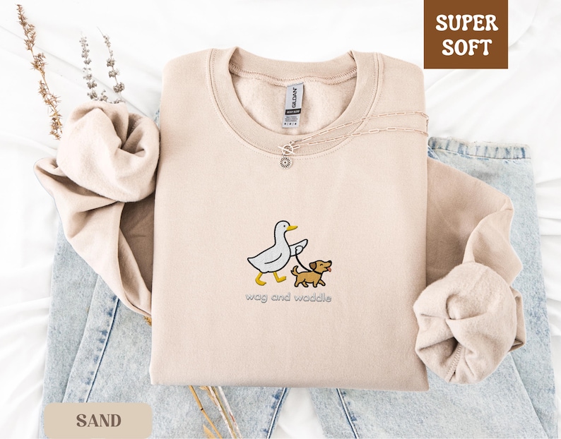 May include: Sand-colored sweatshirt with a crew neck, featuring an embroidered design of a duck walking a dog on a leash. The text "wag and waddle" is below the graphic. The words "Super Soft" are in a brown box in the upper right corner.