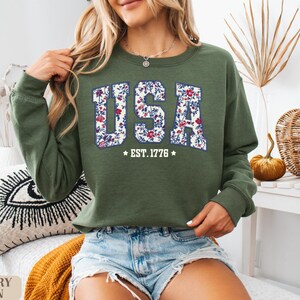 Peut inclure: Sweat-shirt vert olive avec le mot "USA" en motif floral sur la poitrine. Le texte "EST. 1776" est en dessous du mot "USA". Le sweat-shirt est de style col rond.