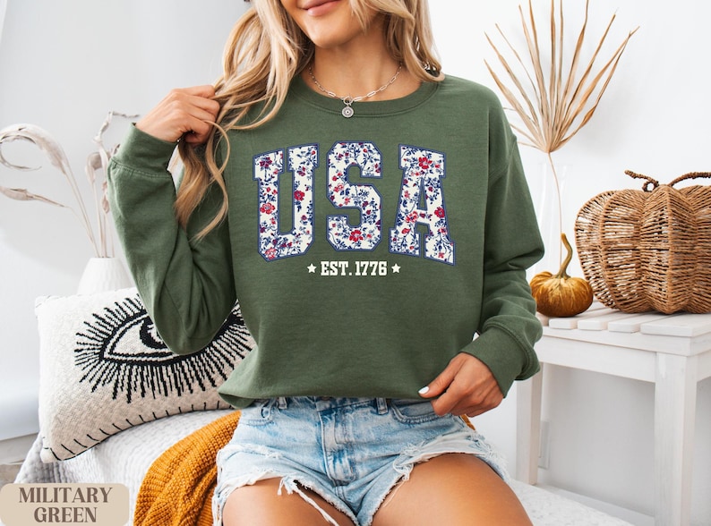 Peut inclure: Sweat-shirt vert olive avec le mot "USA" en motif floral sur la poitrine. Le texte "EST. 1776" est en dessous du mot "USA". Le sweat-shirt est de style col rond.