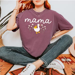 May include: A purple t-shirt with the word "mama" printed in white. A white duck with a yellow chick on its back is printed below the word. The duck is surrounded by small white flowers.
