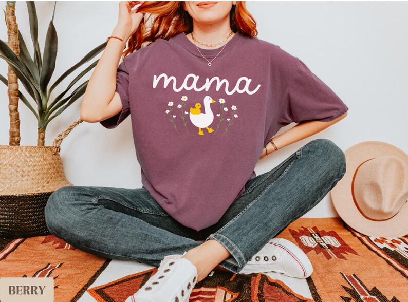 May include: A purple t-shirt with the word "mama" printed in white. A white duck with a yellow chick on its back is printed below the word. The duck is surrounded by small white flowers.