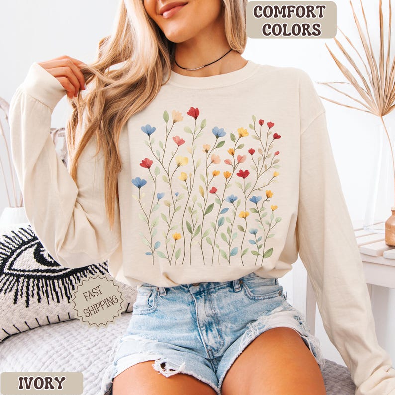 May include: Ivory long-sleeve sweatshirt with a watercolor floral design in red, blue, and yellow. The sweatshirt has the words "Comfort Colors" printed at the top. The word "Ivory" is printed at the bottom.