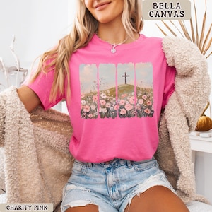 May include: A bright pink t-shirt with a graphic of a cross on a hill, surrounded by flowers. The shirt is labeled "BELLA CANVAS" and "CHARITY PINK". The t-shirt is short-sleeved and made of a soft material.