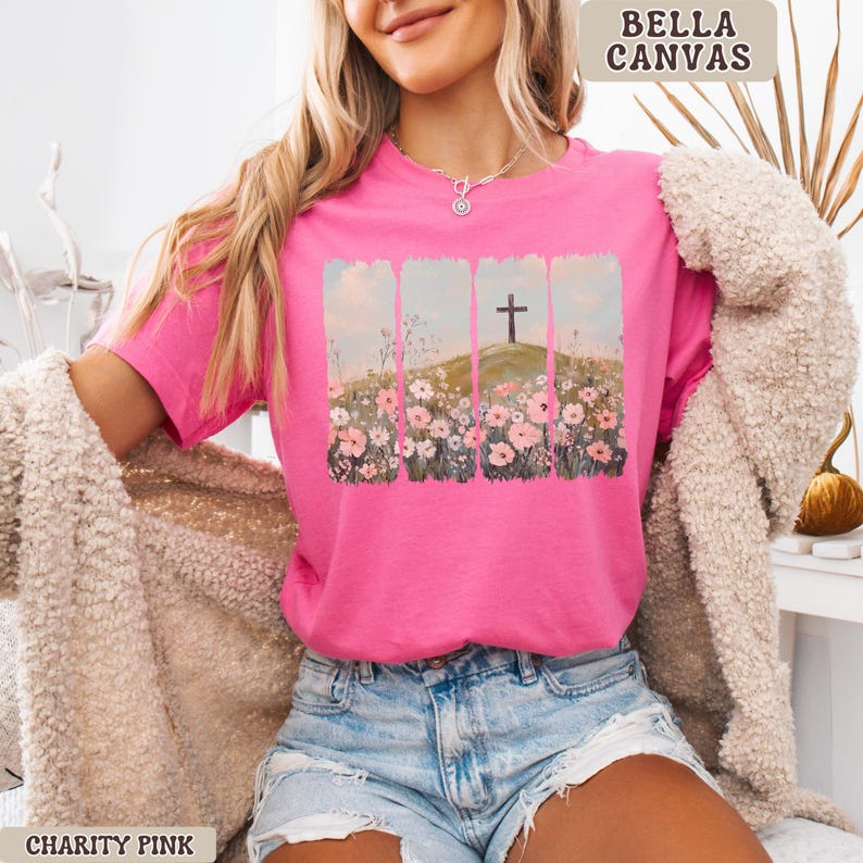 May include: A bright pink t-shirt with a graphic of a cross on a hill, surrounded by flowers. The shirt is labeled "BELLA CANVAS" and "CHARITY PINK". The t-shirt is short-sleeved and made of a soft material.