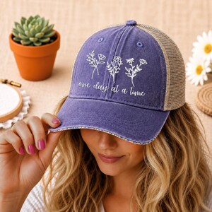 May include: A blue baseball cap with a mesh back and a floral design. The cap features the phrase "one day at a time" in white script. The cap is worn by a person.