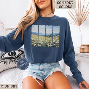 May include: Midnight blue long-sleeve sweatshirt with a landscape painting of a field of white flowers and a blue sky. The words "COMFORT COLORS" are in a tan box at the top right. The word "MIDNIGHT" is at the bottom left.