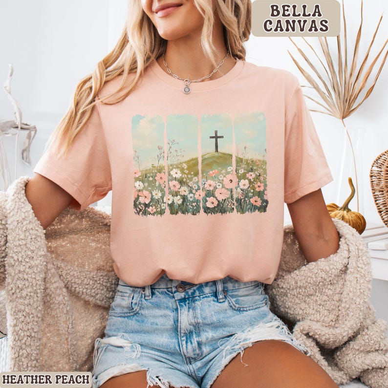 May include: Heather peach-colored t-shirt with a graphic of a cross on a hill, surrounded by flowers. The text "BELLA CANVAS" is in the upper right corner. The text "HEATHER PEACH" is in the lower left corner.