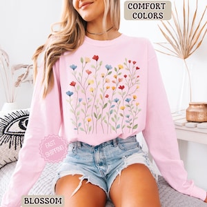 May include: A light pink long-sleeve sweatshirt with a floral design. The design features colorful flowers in red, blue, and yellow. The words "COMFORT COLORS" and "BLOSSOM" are visible.