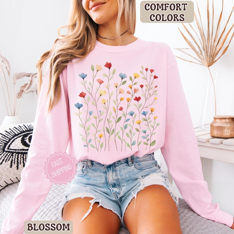 May include: A light pink long-sleeve sweatshirt with a floral design. The design features colorful flowers in red, blue, and yellow. The words "COMFORT COLORS" and "BLOSSOM" are visible.
