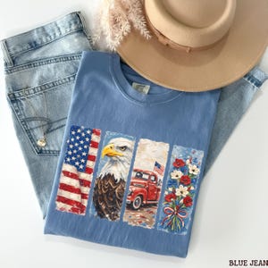 May include: A light blue t-shirt featuring a patriotic design with an American flag, an eagle, a red truck, and flowers. The shirt is paired with light wash blue jeans and a tan hat. A silver necklace is visible.