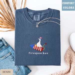 K&ouml;nnte beinhalten: Gefaltetes denimblaues T-Shirt mit einer Grafik einer Gans auf einer Rakete, darunter das Wort "firequacker". Das Shirt hat ein Comfort Colors-Etikett und wird mit einer silbernen Halskette und Jeans kombiniert.