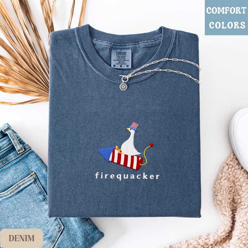K&ouml;nnte beinhalten: Gefaltetes denimblaues T-Shirt mit einer Grafik einer Gans auf einer Rakete, darunter das Wort "firequacker". Das Shirt hat ein Comfort Colors-Etikett und wird mit einer silbernen Halskette und Jeans kombiniert.