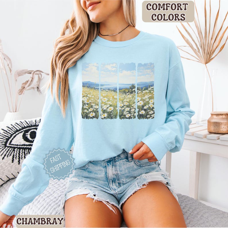 May include: A light blue long-sleeve shirt with a landscape design featuring a field of white daisies under a blue sky. The shirt has the text "Comfort Colors" and "Fast Shipping". The word "Chambray" is also visible.