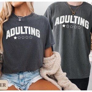 May include: Two dark gray t-shirts with the word "ADULTING" in white, arched text above five stars. The shirts are worn by two people. The shirt on the left is paired with denim shorts. The right shirt is paired with black pants.
