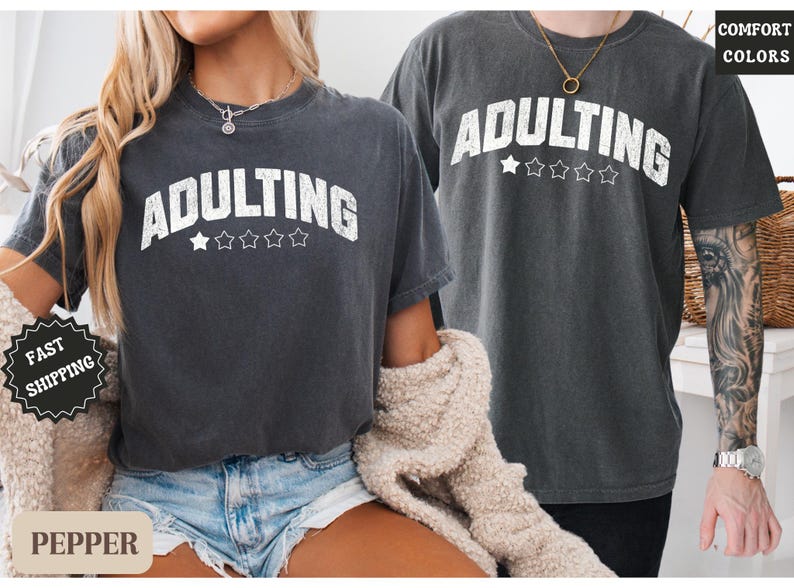 May include: Two dark gray t-shirts with the word "ADULTING" in white, arched text above five stars. The shirts are worn by two people. The shirt on the left is paired with denim shorts. The right shirt is paired with black pants.