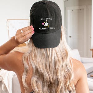 May include: Black baseball cap with white embroidery that reads "Silly Goose Pickleball Club". The embroidery also features a cartoon goose holding a pickleball paddle.