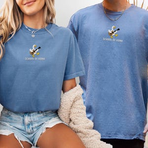 May include: Two blue jean-colored t-shirts. Each shirt has a small embroidered design of a duck playing a guitar, with the text "SCHOOL OF HONK" below. The shirts are short-sleeved and have a relaxed fit.
