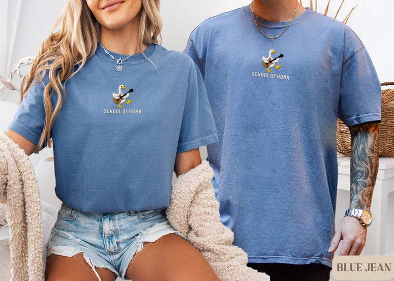 May include: Two blue jean-colored t-shirts. Each shirt has a small embroidered design of a duck playing a guitar, with the text "SCHOOL OF HONK" below. The shirts are short-sleeved and have a relaxed fit.