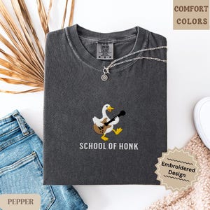 May include: Dark gray t-shirt with an embroidered design of a goose playing a guitar. The text "SCHOOL OF HONK" is below the goose. A silver necklace is draped across the shirt. The shirt is folded on a white surface.