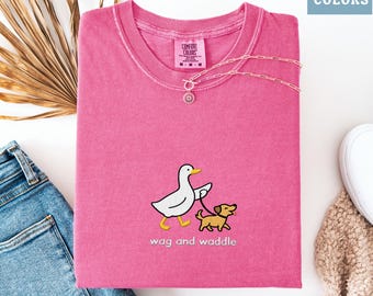 May include: A folded, bright pink t-shirt with a graphic of a duck walking a dog on a leash, with the words "wag and waddle" below. A silver necklace with a circular pendant rests on the shirt. The shirt is labeled "Comfort Colors".