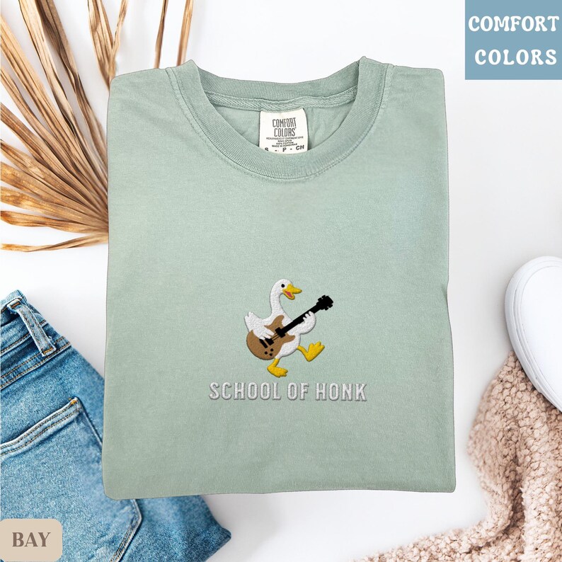 May include: A light green sweatshirt with the text "SCHOOL OF HONK" and an embroidered duck playing a guitar. The sweatshirt is folded and placed on a white surface with a pair of blue jeans.