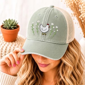 May include: A sage green trucker hat with a white chicken and floral embroidery. The hat has a mesh back and a curved brim with a white trim. The hat is a casual accessory.