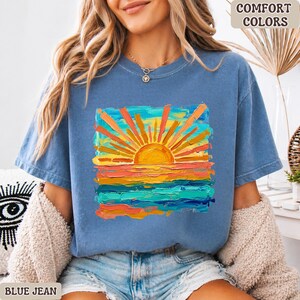 Puede incluir: Camiseta azul vaquero con un gr&aacute;fico de puesta de sol colorido. El dise&ntilde;o presenta un sol vibrante con rayos naranjas sobre un mar de pinceladas azules, naranjas y amarillas. Las palabras "COMFORT COLORS" est&aacute;n impresas en la parte superior derecha.