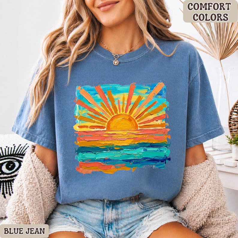 Puede incluir: Camiseta azul vaquero con un gr&aacute;fico de puesta de sol colorido. El dise&ntilde;o presenta un sol vibrante con rayos naranjas sobre un mar de pinceladas azules, naranjas y amarillas. Las palabras "COMFORT COLORS" est&aacute;n impresas en la parte superior derecha.
