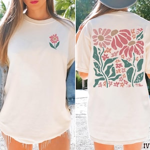 May include: Ivory-colored t-shirt featuring a floral design. The front displays a single pink flower with green leaves, while the back showcases a larger floral arrangement in pink, brown, and green. The shirt has a crew neck and short sleeves.