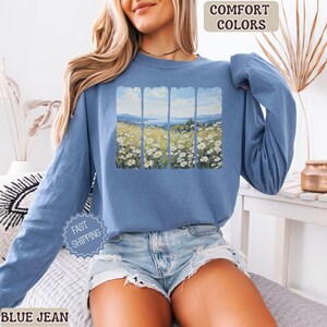May include: A blue long-sleeve sweatshirt with a landscape graphic. The graphic depicts a field of white flowers, a body of water, and a blue sky. The text "COMFORT COLORS" is at the top. The sweatshirt is paired with denim shorts.