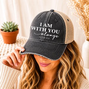 May include: A black and tan trucker hat with the text "I AM WITH YOU always" in white script. The hat has a curved brim and a mesh back. The hat is a casual accessory.