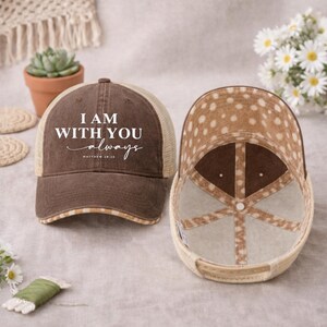 May include: Brown and tan trucker hat with the text "I AM WITH YOU always" in white script. The hat has a mesh back and a patterned interior. The hat is displayed next to another hat with the interior facing up.