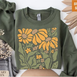 May include: Military green sweatshirt with a floral design featuring large yellow and orange flowers, green leaves, and small brown and yellow flowers. The words "SUPER SOFT" are in the upper right corner.