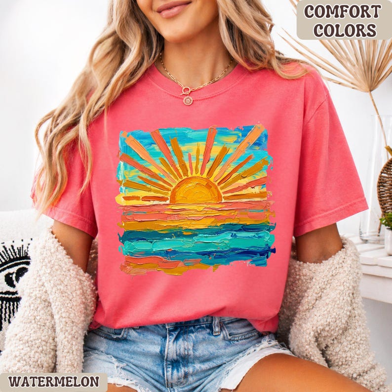 Puede incluir: Una camiseta de color coral con un vibrante gr&aacute;fico de puesta de sol. La obra de arte presenta un sol amarillo brillante con rayos naranjas y amarillos radiantes sobre un oc&eacute;ano colorido. Las palabras "COMFORT COLORS" y "WATERMELON" son visibles.