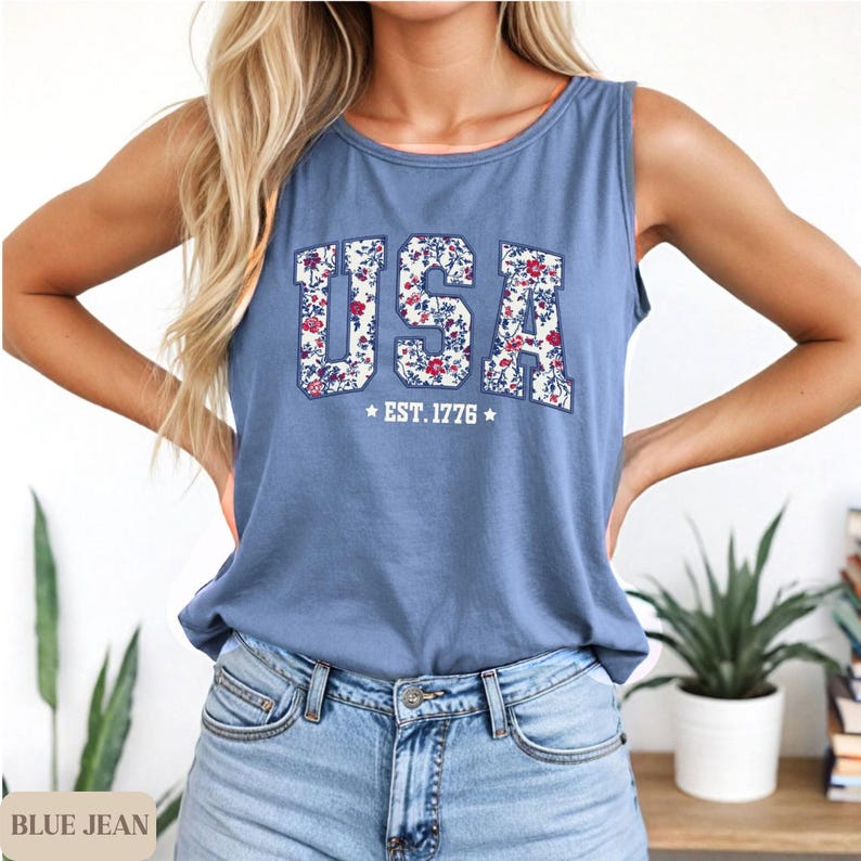 May include: A blue tank top with the word "USA" in a floral design. The text "EST. 1776" is below the word "USA". The tank top is sleeveless and made of a soft material. The tank top is paired with blue jeans.