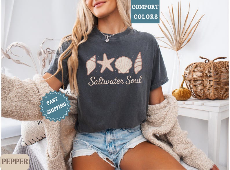 May include: Dark gray t-shirt with a seashell, starfish, and seashell design, and the words "Saltwater Soul". The shirt is a comfortable fit and is perfect for a beach or ocean theme.
