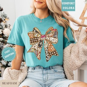 May include: Seafoam green t-shirt featuring a large patchwork bow design. The bow incorporates various floral and plaid patterns in shades of brown, orange, and white. The shirt has short sleeves and a crew neck. The words "Comfort Colors" are printed on the top right.