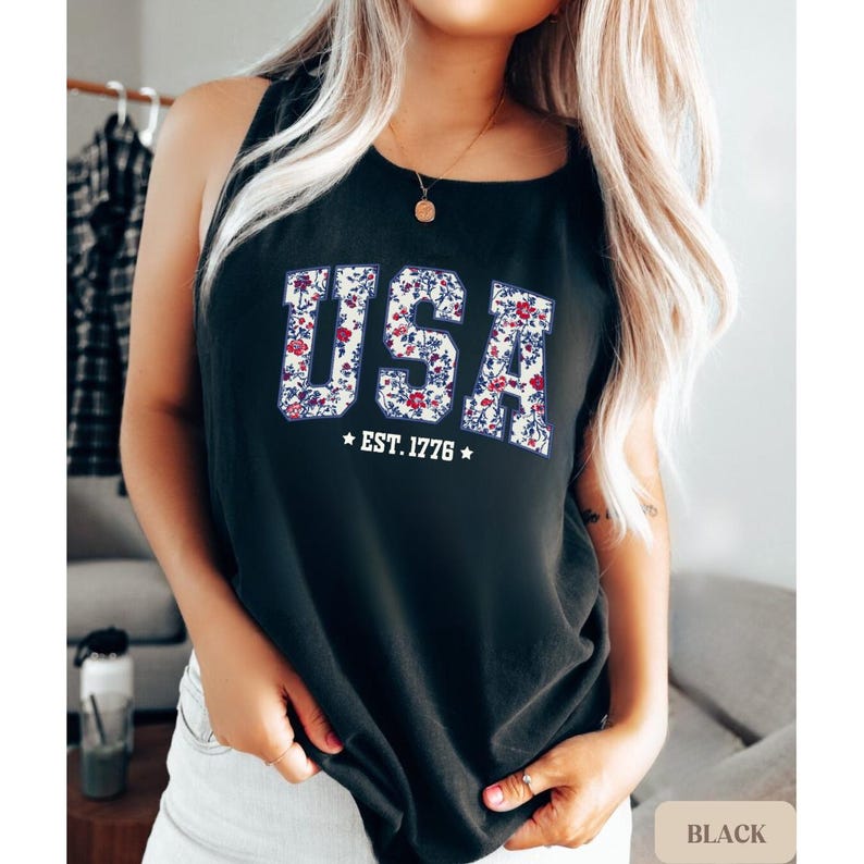 K&ouml;nnte beinhalten: Schwarzes Tanktop mit dem Wort "USA" in wei&szlig;en, bl&uuml;tengef&uuml;llten Buchstaben. Darunter steht der Text "EST. 1776" mit Sternen auf beiden Seiten. Das Tanktop ist &auml;rmellos.