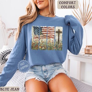 Op de afbeelding: Blauw shirt met lange mouwen met een afbeelding van de Amerikaanse vlag en een houten kruis. Het ontwerp bevat een bloemenweide. De tekst "Comfort Colors" en "Blue Jean" is ook aanwezig.