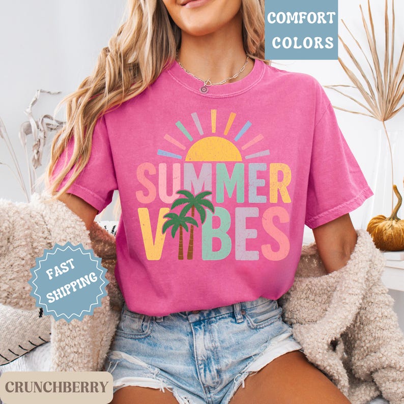 K&ouml;nnte beinhalten: Ein pinkfarbenes T-Shirt mit den Worten "SUMMER VIBES" in bunten Buchstaben, einer Sonne und Palmen. Das Shirt hat kurze &Auml;rmel und wird mit Jeansshorts getragen. Die Worte "COMFORT COLORS" und "FAST SHIPPING" sind ebenfalls sichtbar.