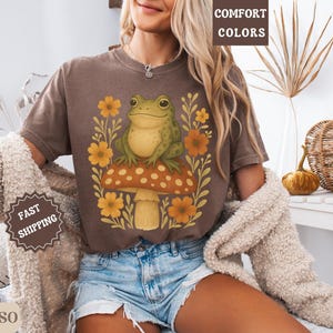 May include: A brown t-shirt featuring a graphic of a green frog perched on a mushroom with flowers. The text "COMFORT COLORS" is visible in the top right corner of the image.