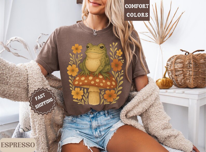 May include: A brown t-shirt featuring a graphic of a green frog perched on a mushroom with flowers. The text "COMFORT COLORS" is visible in the top right corner of the image.