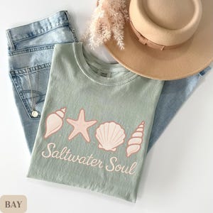 May include: A sage green t-shirt with a seashell design and the words "Saltwater Soul" in a script font. The shirt is paired with light wash denim jeans and a tan hat.