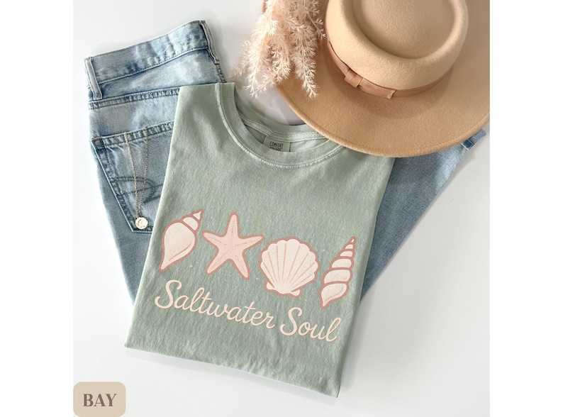May include: A sage green t-shirt with a seashell design and the words "Saltwater Soul" in a script font. The shirt is paired with light wash denim jeans and a tan hat.