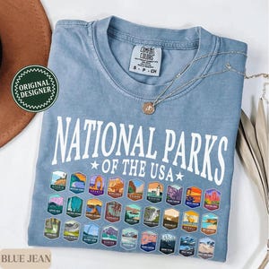 以下が含まれることがあります： 薄い青色のTシャツに、白い文字で「National Parks of the USA」と書いてあり、カラフルな国立公園のロゴが並んでいます。