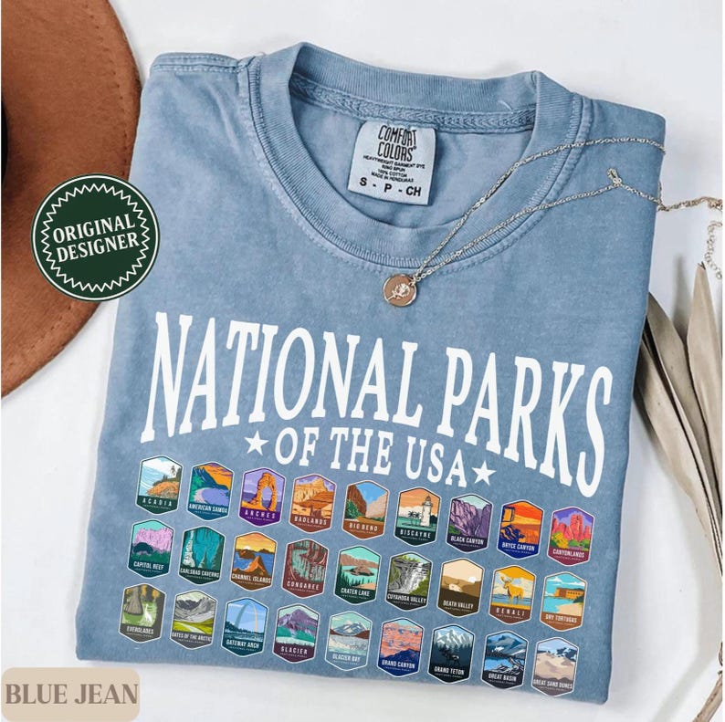 以下が含まれることがあります： 薄い青色のTシャツに、白い文字で「National Parks of the USA」と書いてあり、カラフルな国立公園のロゴが並んでいます。
