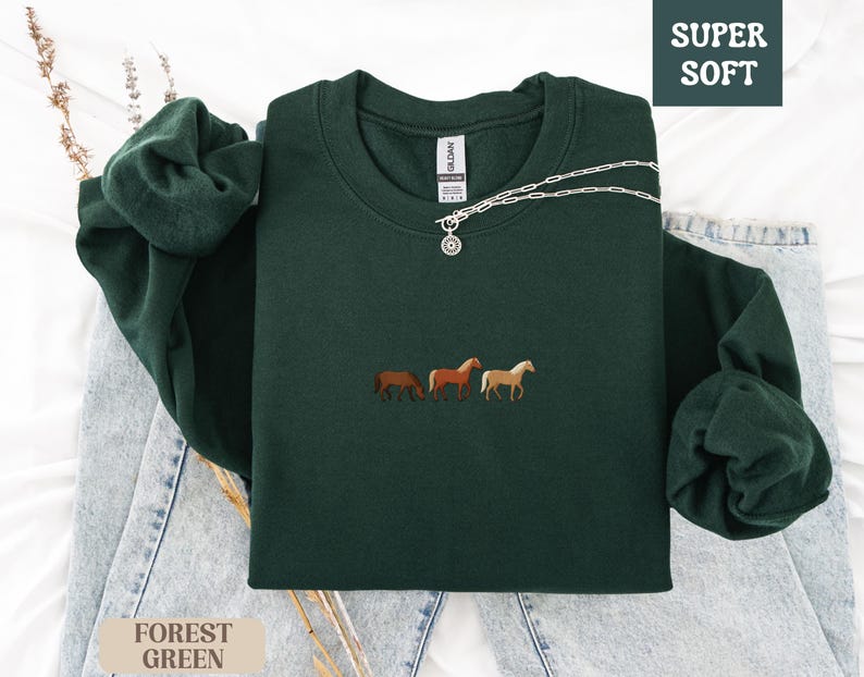 K&ouml;nnte beinhalten: Dunkelgr&uuml;nes Sweatshirt mit gestickten Pferden. Auf der rechten oberen Ecke des Sweatshirts steht &bdquo;SUPER SOFT&ldquo;. Eine silberne Halskette mit rundem Anh&auml;nger liegt &uuml;ber der Vorderseite. Das Sweatshirt ist auf einer Jeans gefaltet.