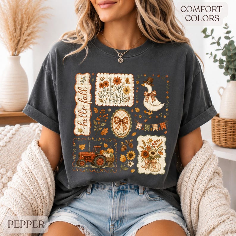 May include: A charcoal gray short-sleeve t-shirt featuring a graphic print with the text "hello fall" and various autumn-themed illustrations including a tractor, a goose wearing a hat, sunflowers, pumpkins, and a plaid bow.